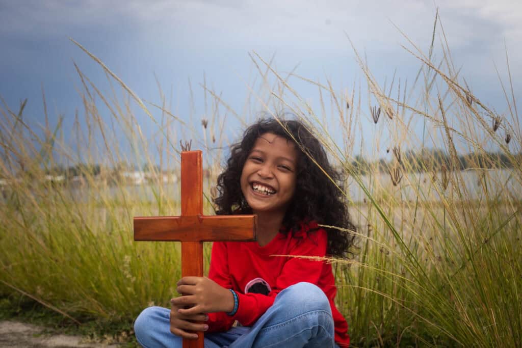 A young girl sits among tall grass while holding a wooden cross and smiling for the camera.