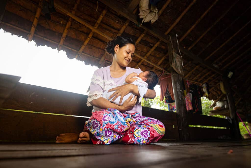 A mother sits on the ground holding her baby. She looks down smiling at her child.