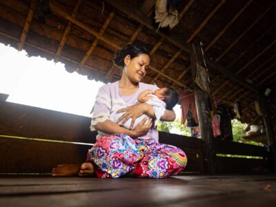 A mother sits on the ground holding her baby. She looks down smiling at her child.