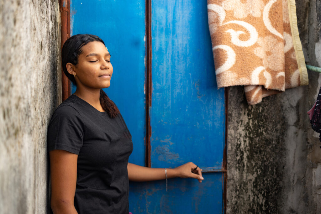 A young girl leans against a wall with her eyes closed.