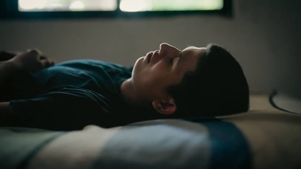 A young man lies on a bed in the dark with his eyes closed.