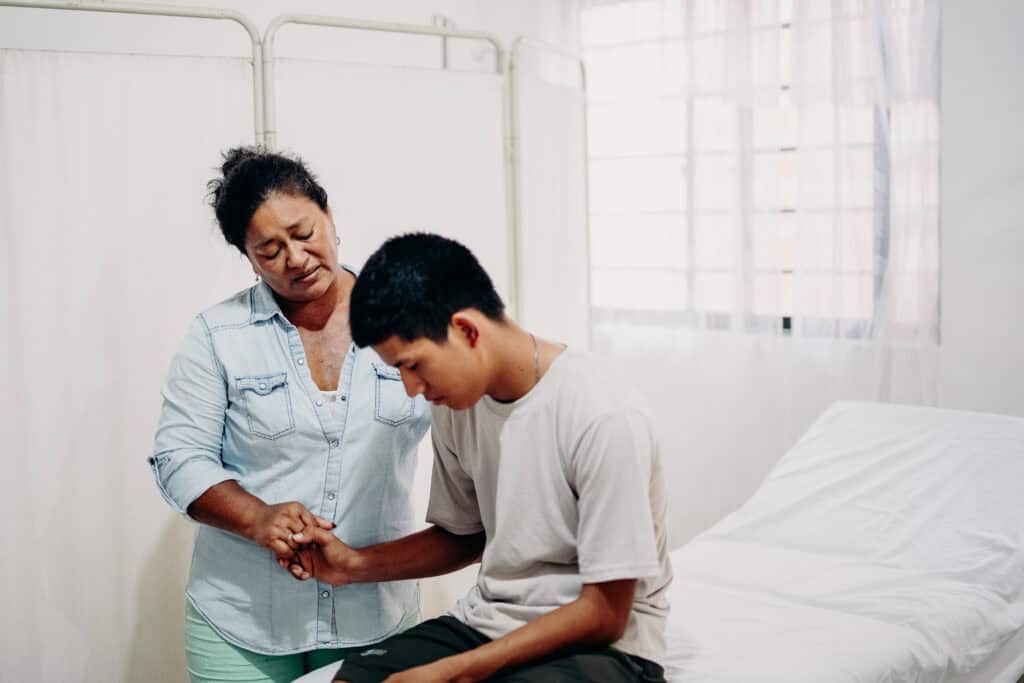 A young man sits on a hospital bed holding the hand of an older woman as they pray together.