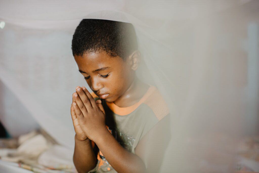 A young boy sits with his eyes closed and hands together in front of his face.