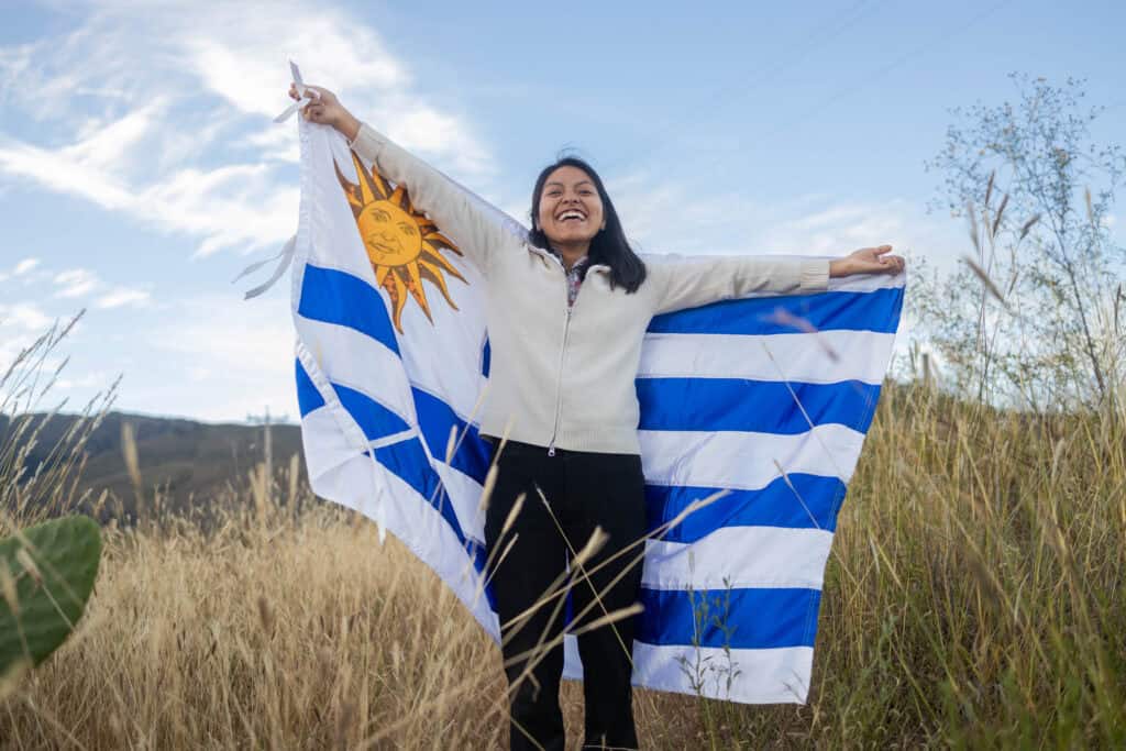 A young woman stands in a field holding the flag of Uruguay while smiling.