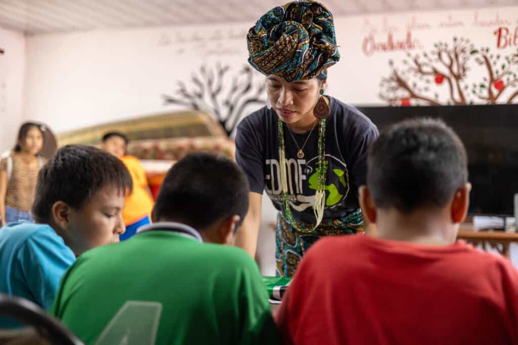 A woman stands in front of 3 children sitting at a desk. 