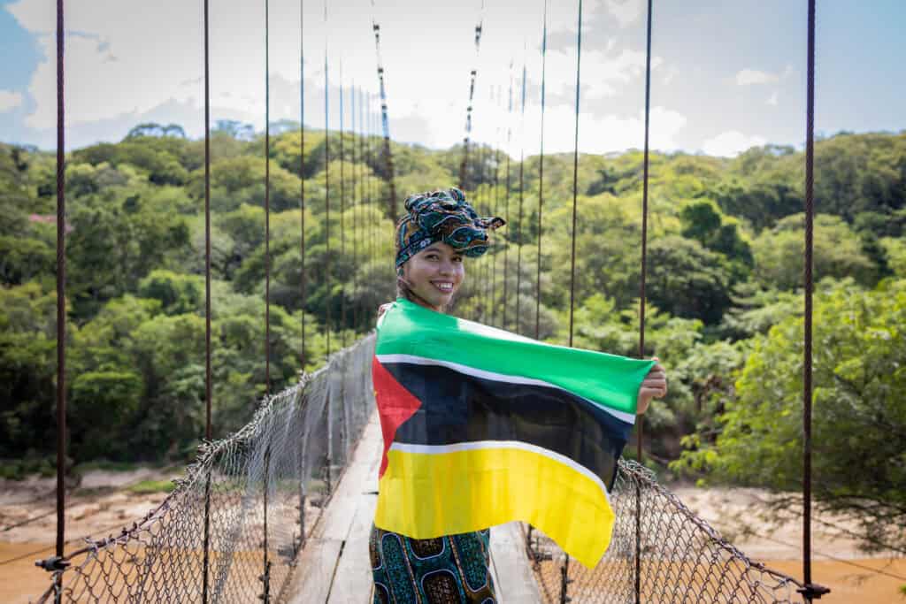 A young woman wearing a headscarf stands on a bridge holding the flag of Mozambique.