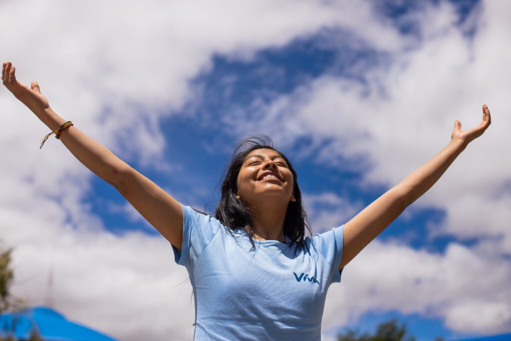 A young girl wearing a blue shirt raises her arms in front of a cloud-filled blue sky.