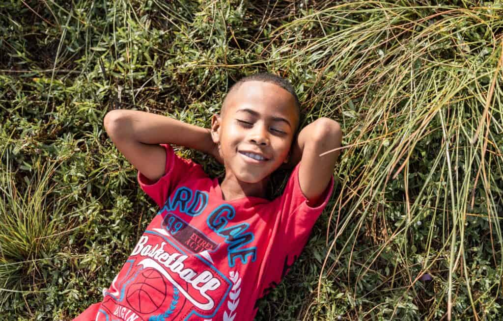 A young boy lies on the grass with his hands behind his head and his eyes closed.