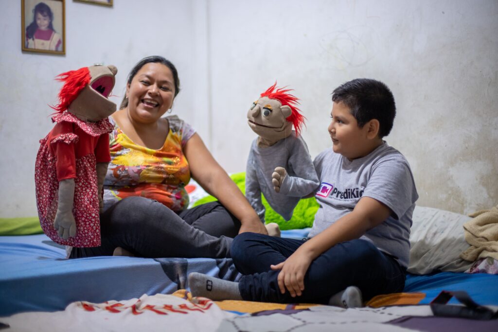 A young boy and an older woman hold puppets while sitting on a bed.