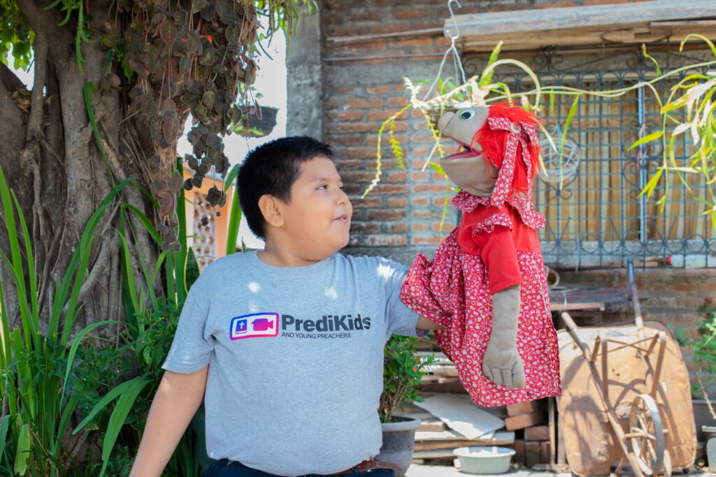 A young boy holds a puppet wearing a red patterned dress.