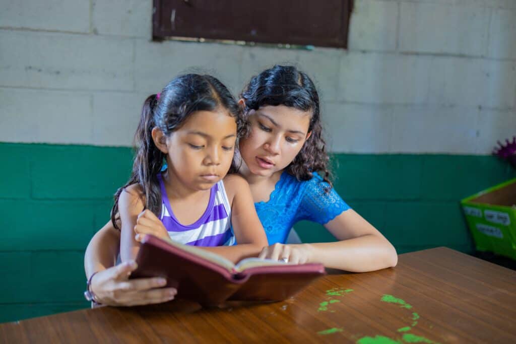 Two young girls sit together while one girl reads the Bible to the other.