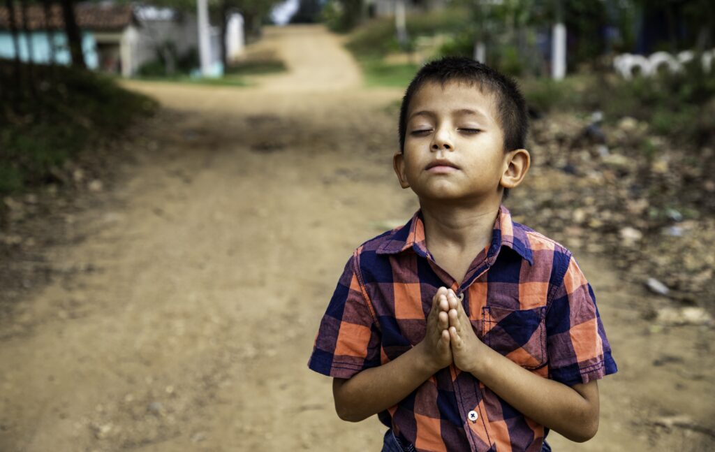 A young boy wearing a plaid shirt clasps his hands together prays with his eyes closed.