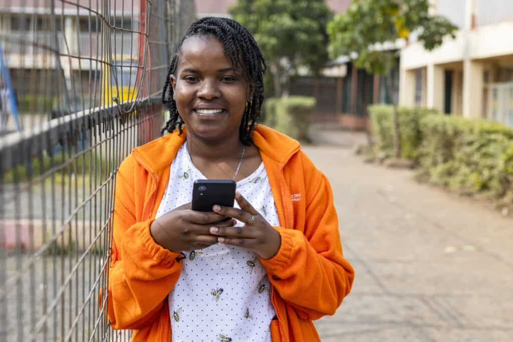 A girl wearing a bright orange jacket smiles for the camera while holding a cell phone.