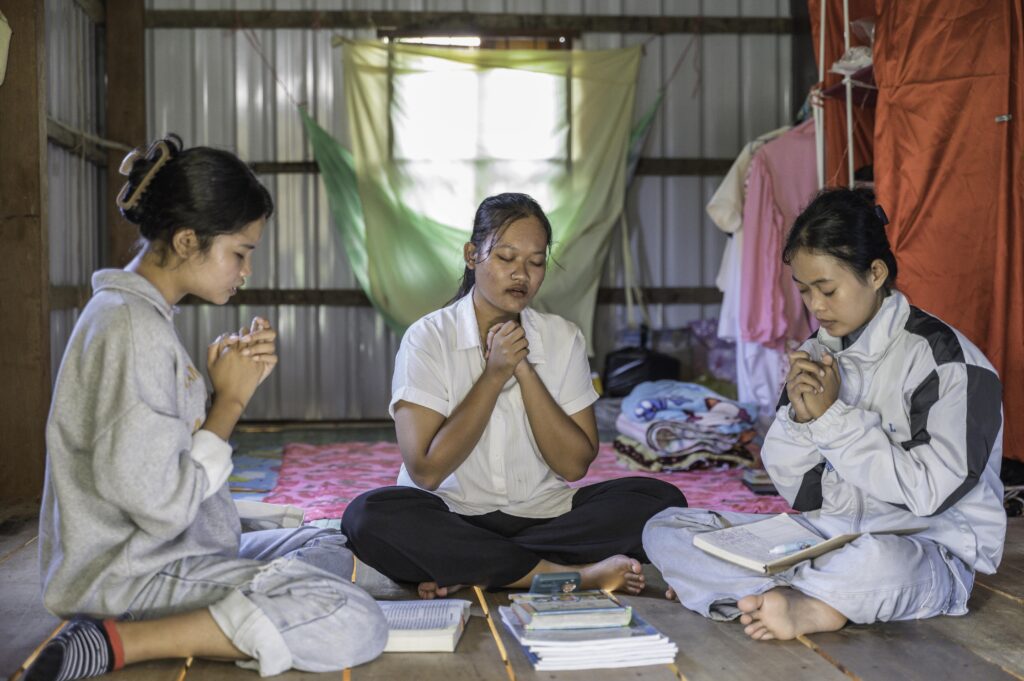 Three young women sit in a circle with their hands clasped in front of them and their eyes closed.