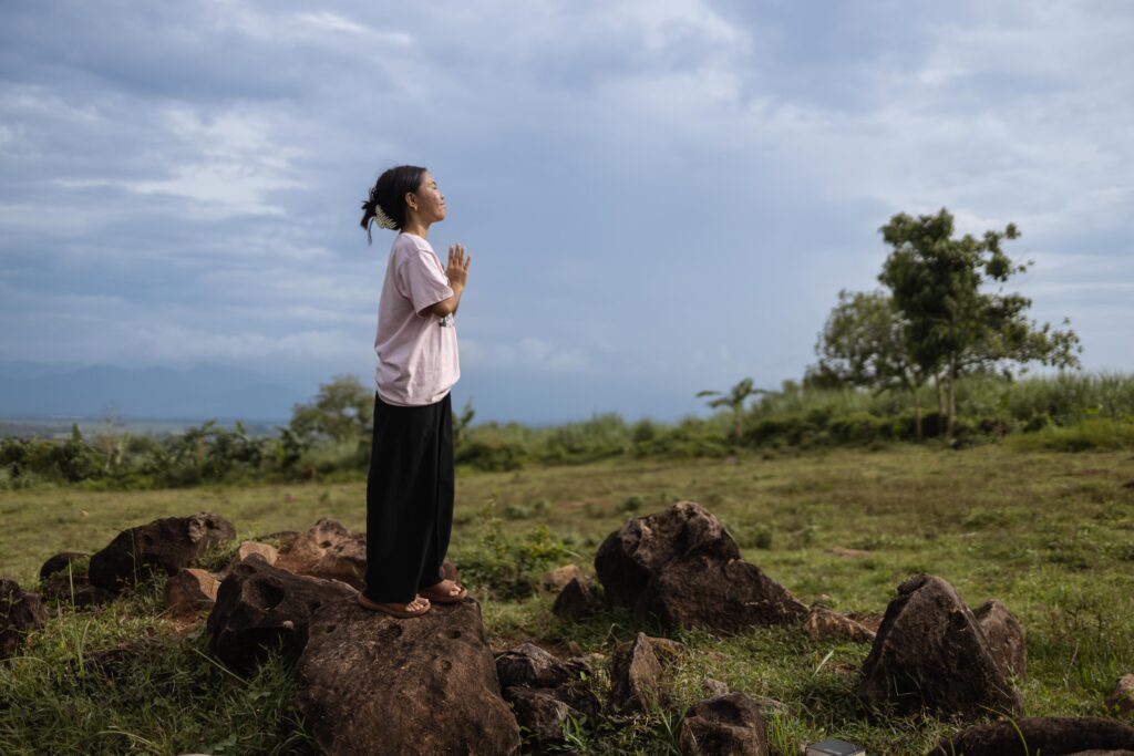 A young woman stands on a rock with her hands pressed together and her head lifted to the sky.