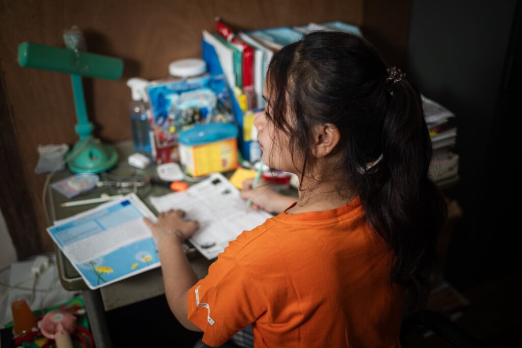 A young girl wearing an orange shirt sits at a desk and looks to the left.