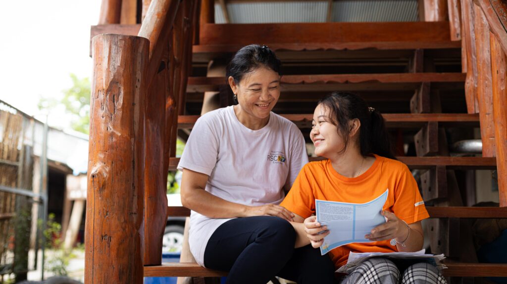 A young girl wearing a bright orange shirt sits with an older woman on steps while holding a letter and smiling.