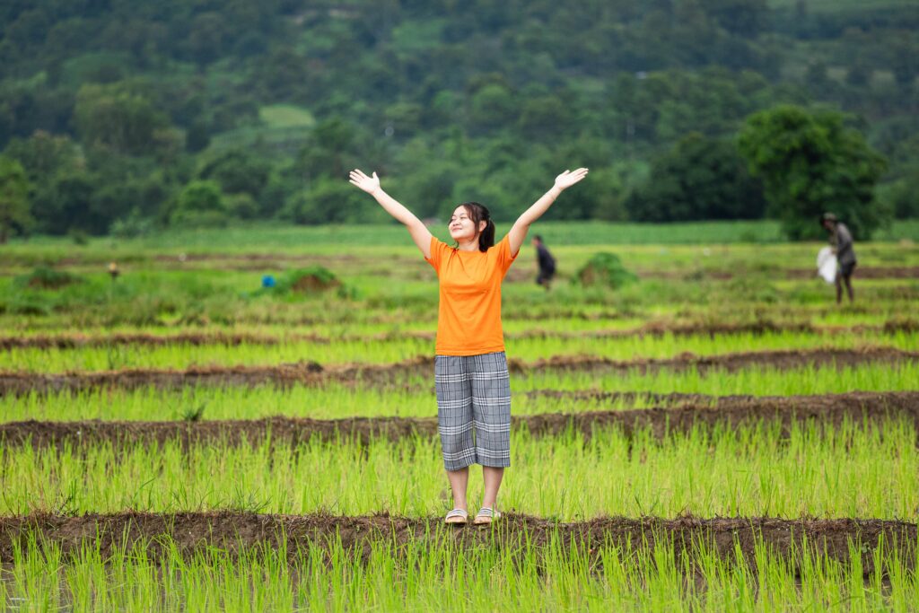 A girl wearing a bright orange shirt stands in a field with her arms stretched to the sky.