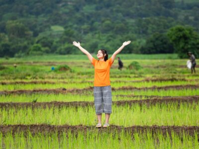 A girl wearing a bright orange shirt stands in a field with her arms stretched to the sky.