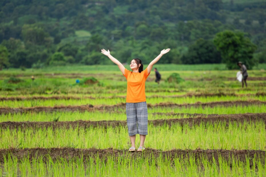 A girl wearing a bright orange shirt stands in a field with her arms stretched to the sky.