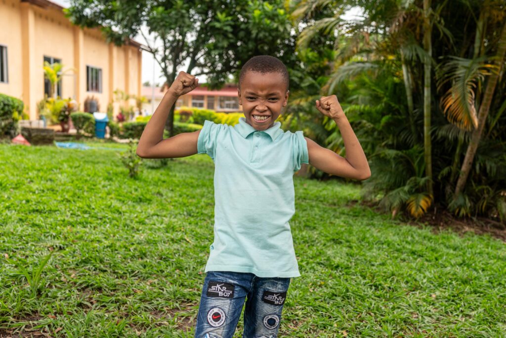 A young boy wearing a mint green polo flexes his arms in front of the camera.