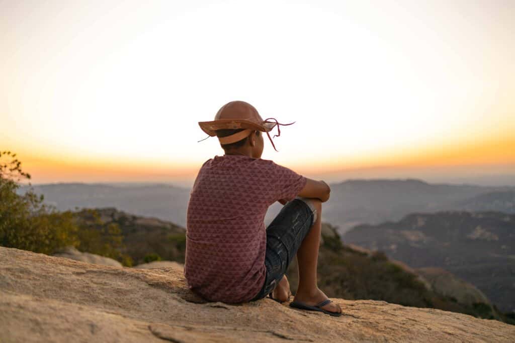 A young man wearing a hat sits on the ground with his back turned to the camera. The sunset shines in front of him.