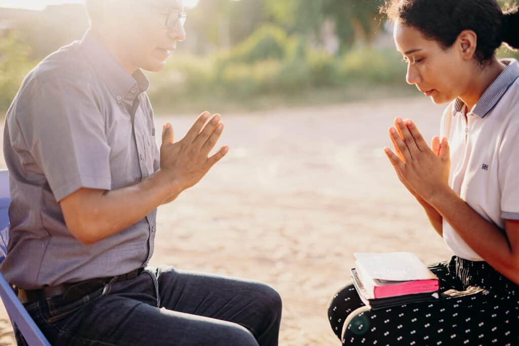 A young woman sits with her hands clasped together and a Bible in her lap. An older man sits in front of her and prays.