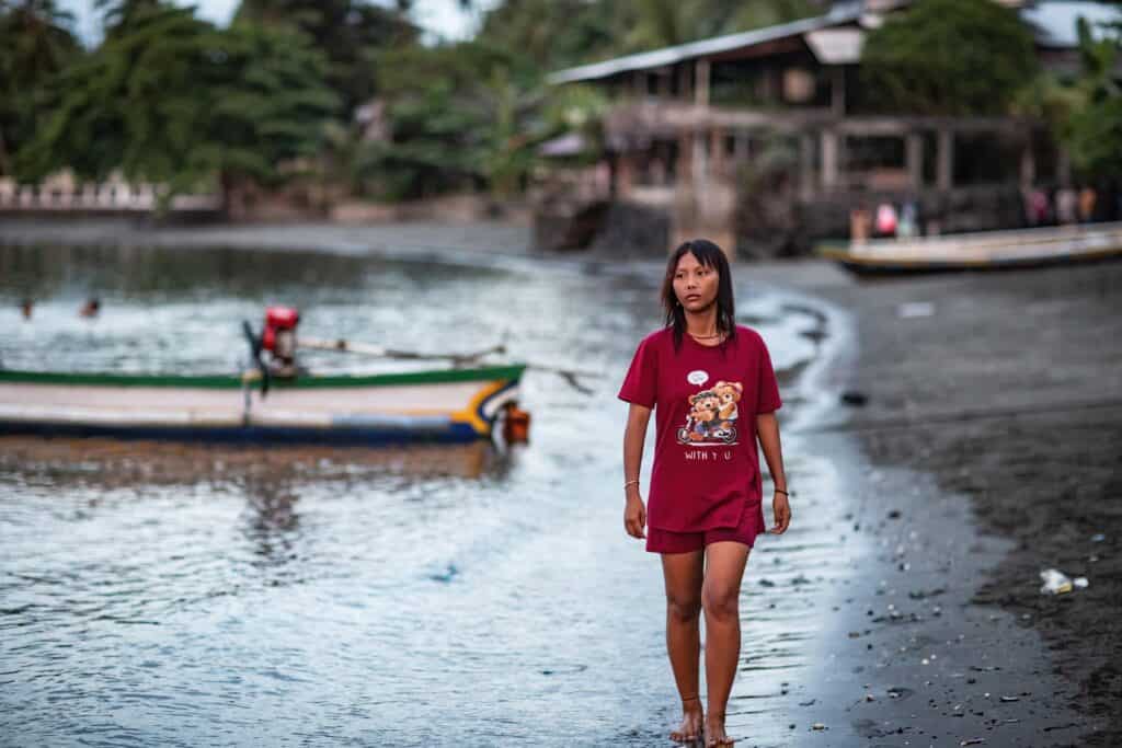 A young girl wearing a red shirt and shorts walks along the shore. A boat is in the background.