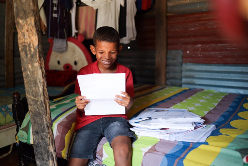 A young boy sits on a bed while reading a letter and smiling.