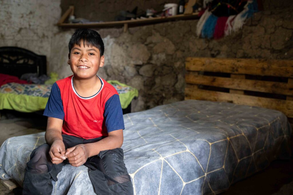 A young boy sits on a bed while smiling for the camera.