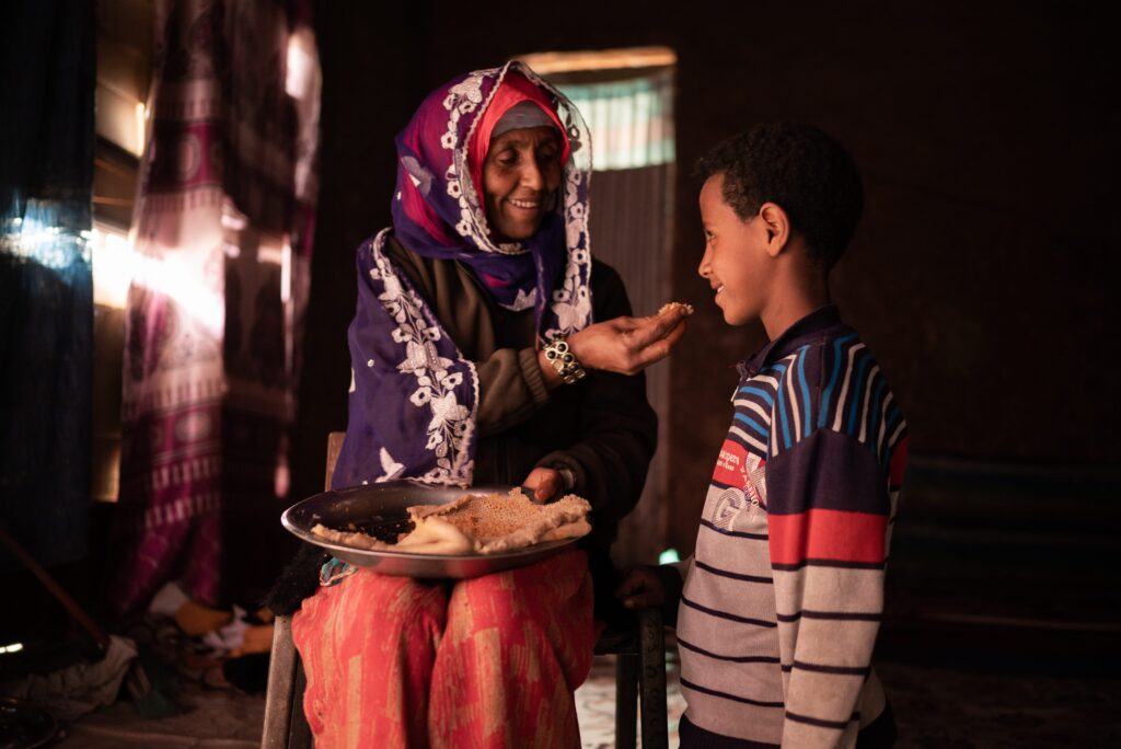 A woman wearing a head covering feeds a little boy as they smile at each other.