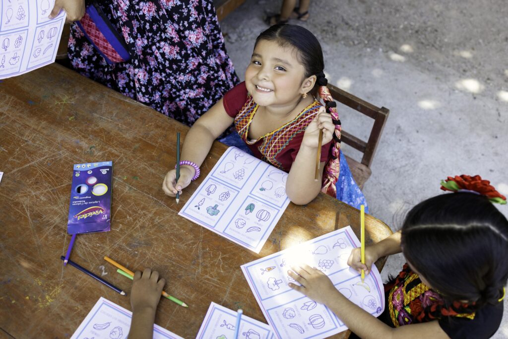 A little girl sits in a wooden chair at a wooden table while coloring a picture of vegetables.