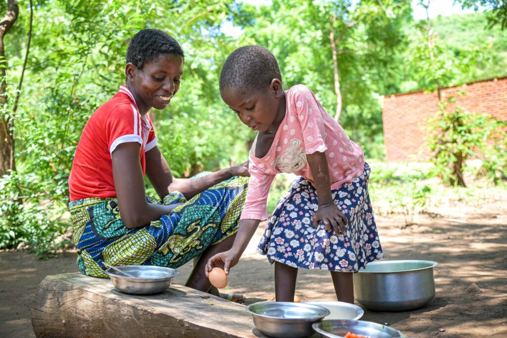 A little girl cracks open an egg while her mother sits and smiles next to her.