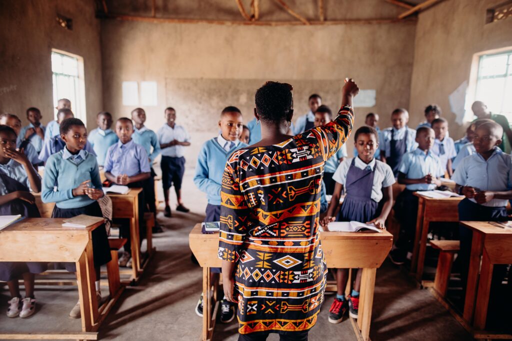 A woman wearing a brightly patterned dress stands in front of a classroom of children wearing school uniforms.