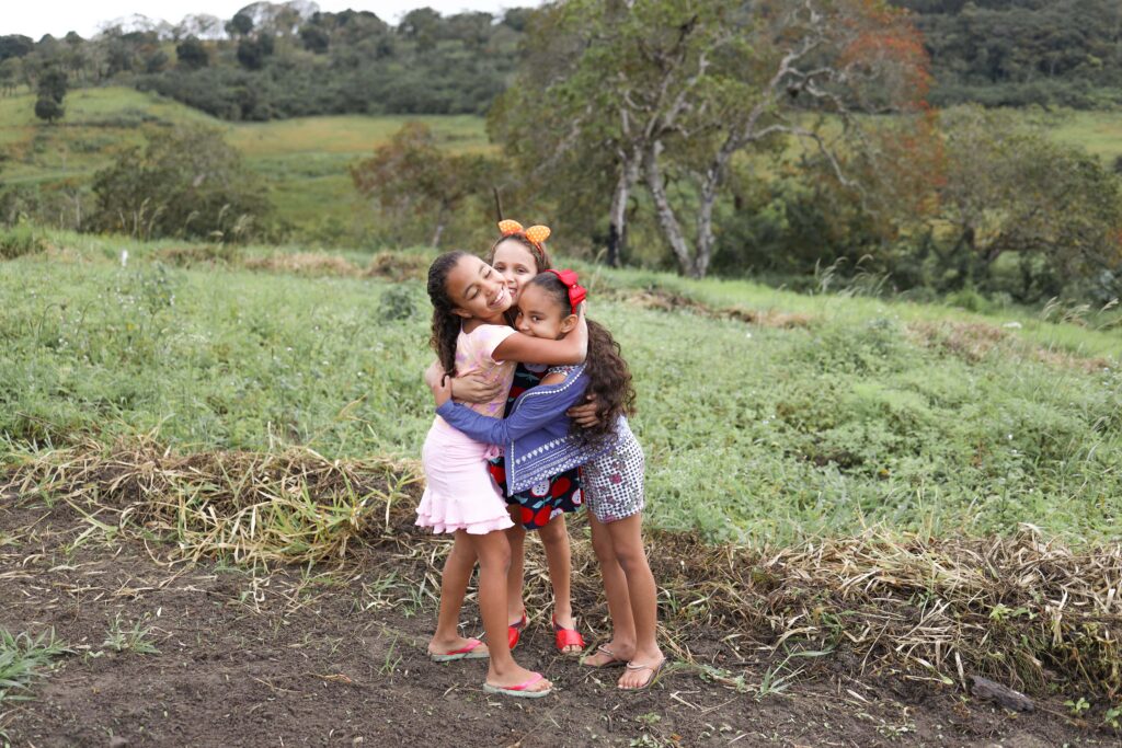 Three young girls hug each other in front of a lush green landscape.