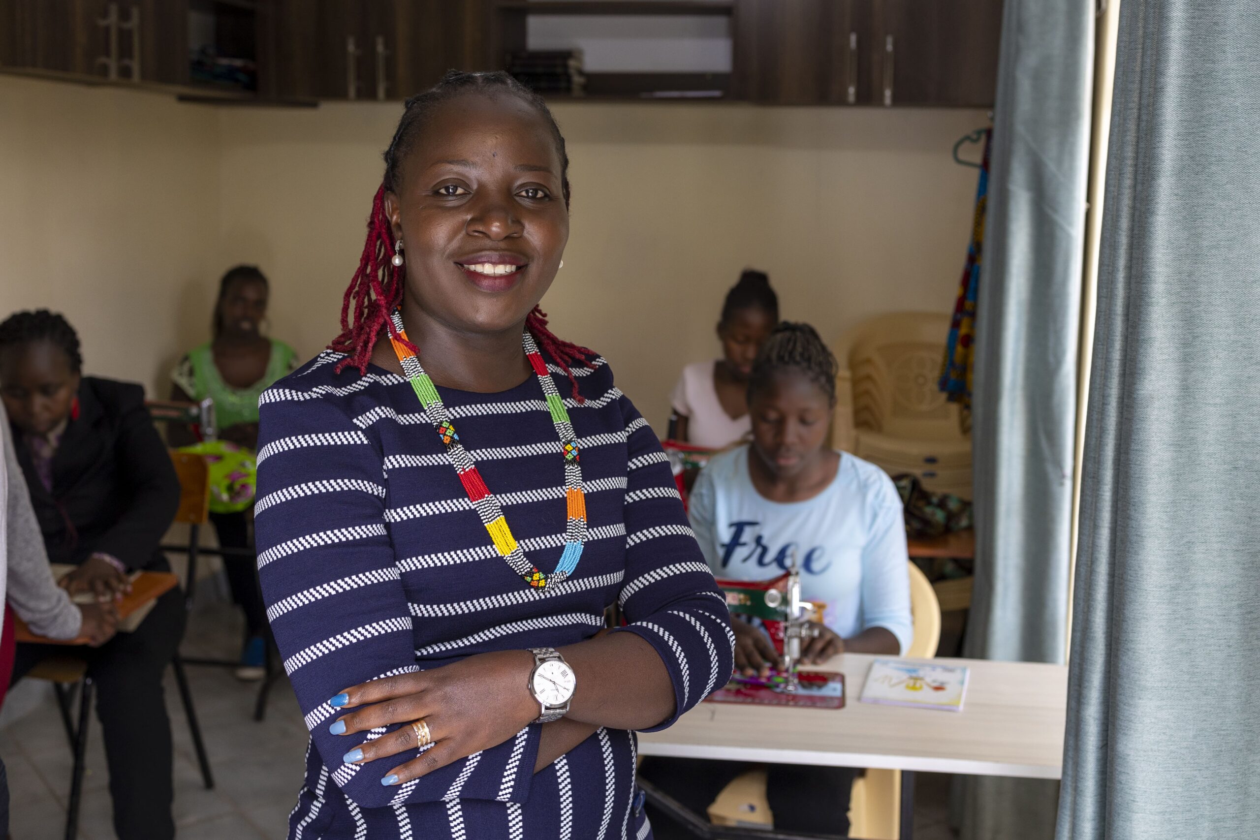 CC-KE0909-alumni-2210-8-min An older woman wearing a blue and white striped dress crosses her arms and smiles for the camera in front of a classroom of teen girls.