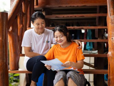 CC-TH095400201-08-2308-min A young girl wearing an orange shirt reads a letter next to an older woman while smiling.