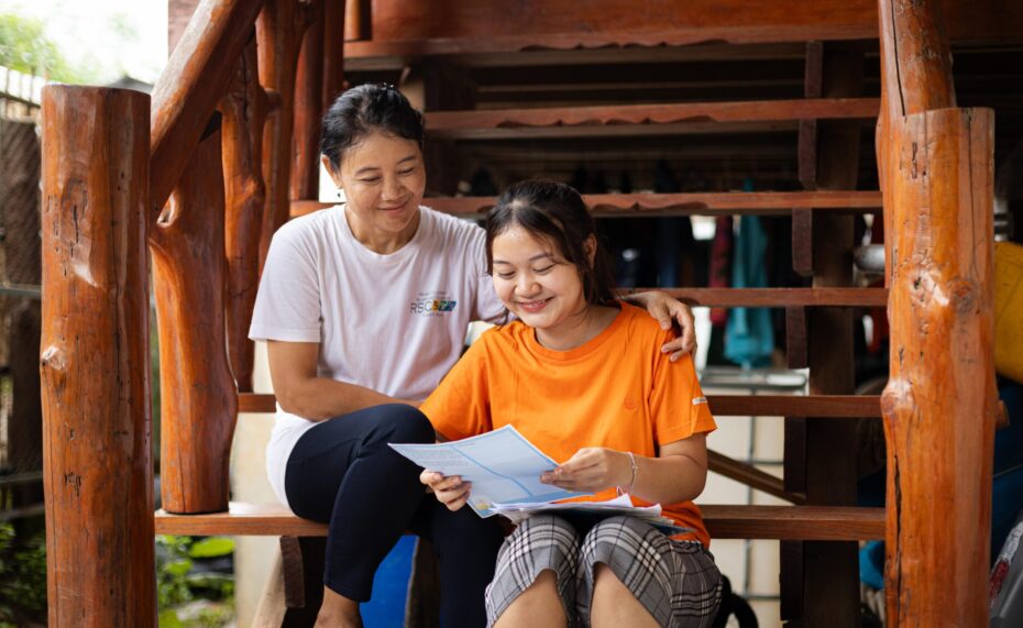 CC-TH095400201-08-2308-min A young girl wearing an orange shirt reads a letter next to an older woman while smiling.