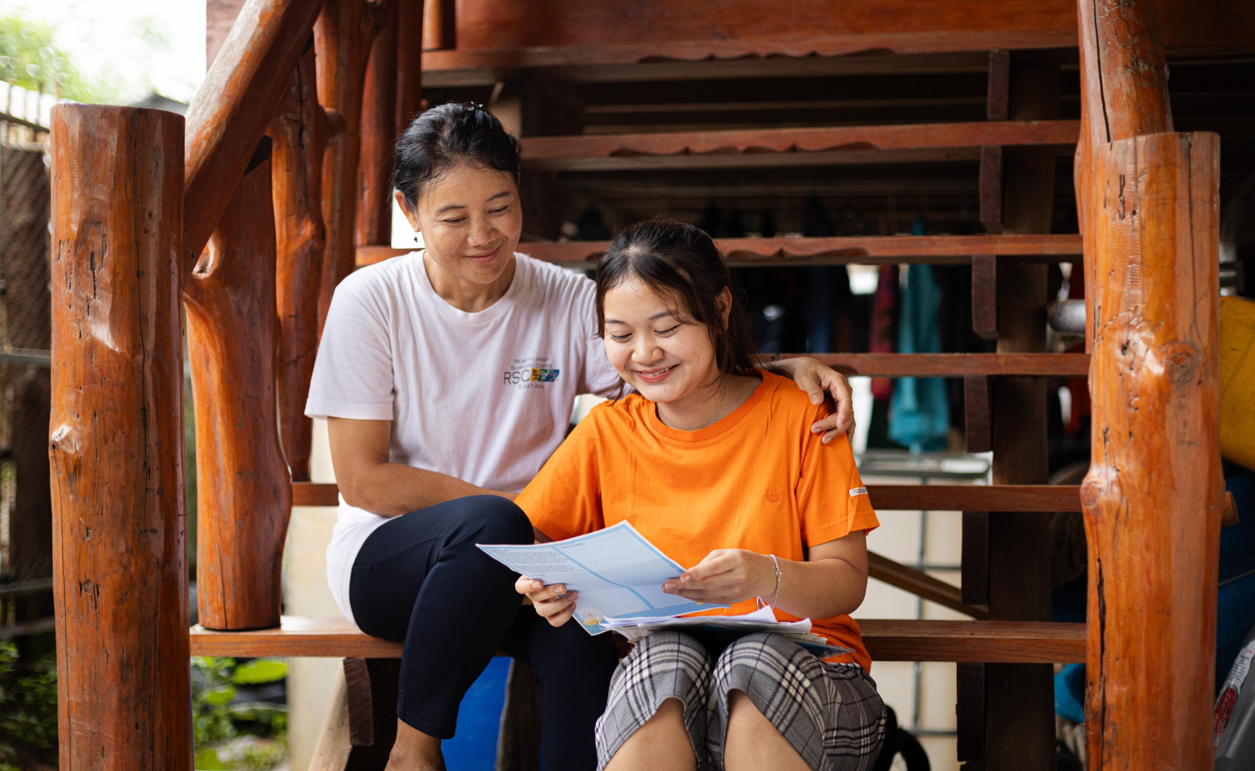 CC-TH095400201-08-2308-min A young girl wearing an orange shirt reads a letter next to an older woman while smiling.