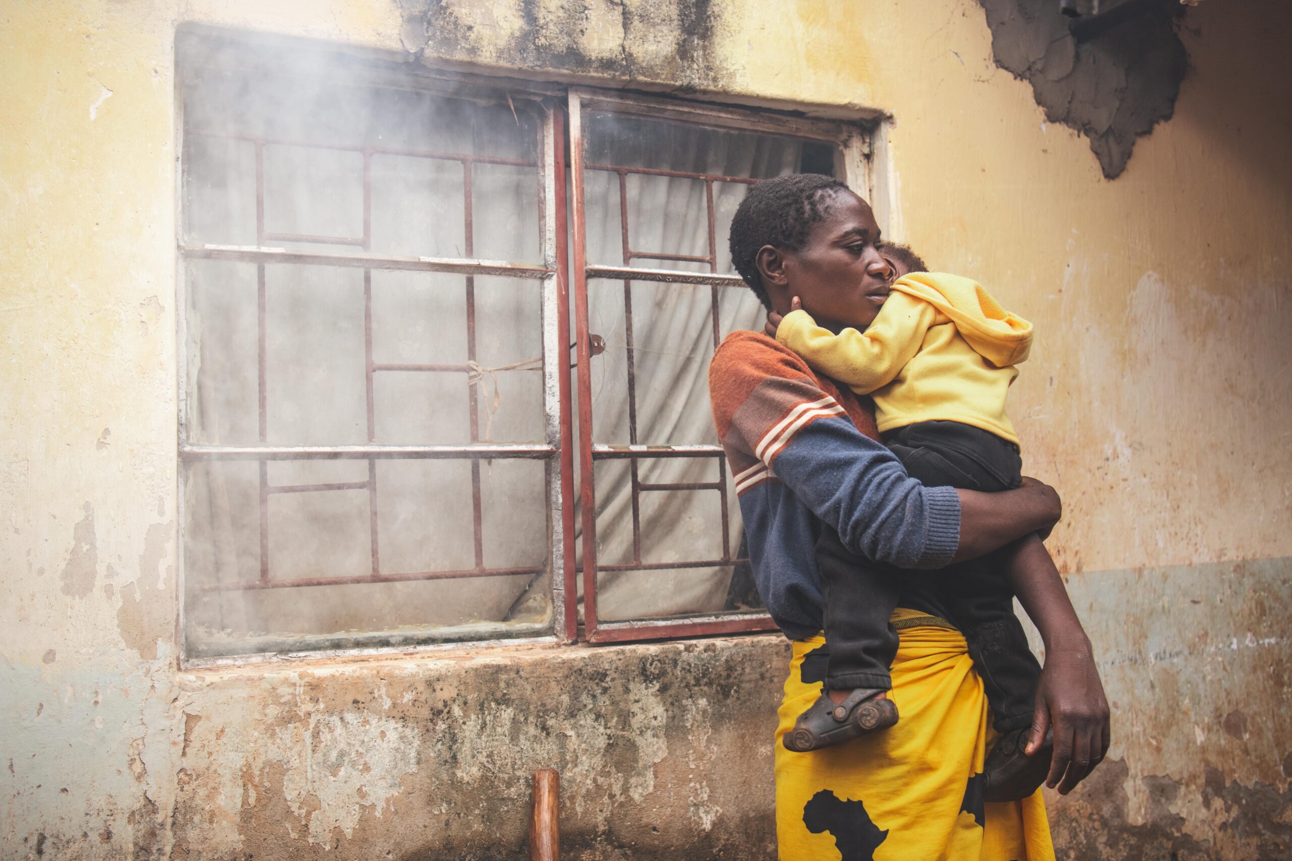 CC-ZM0104-NCL-From Ashes to Beauty -2502-12-min A young woman holds her toddler child while standing in front of a window.