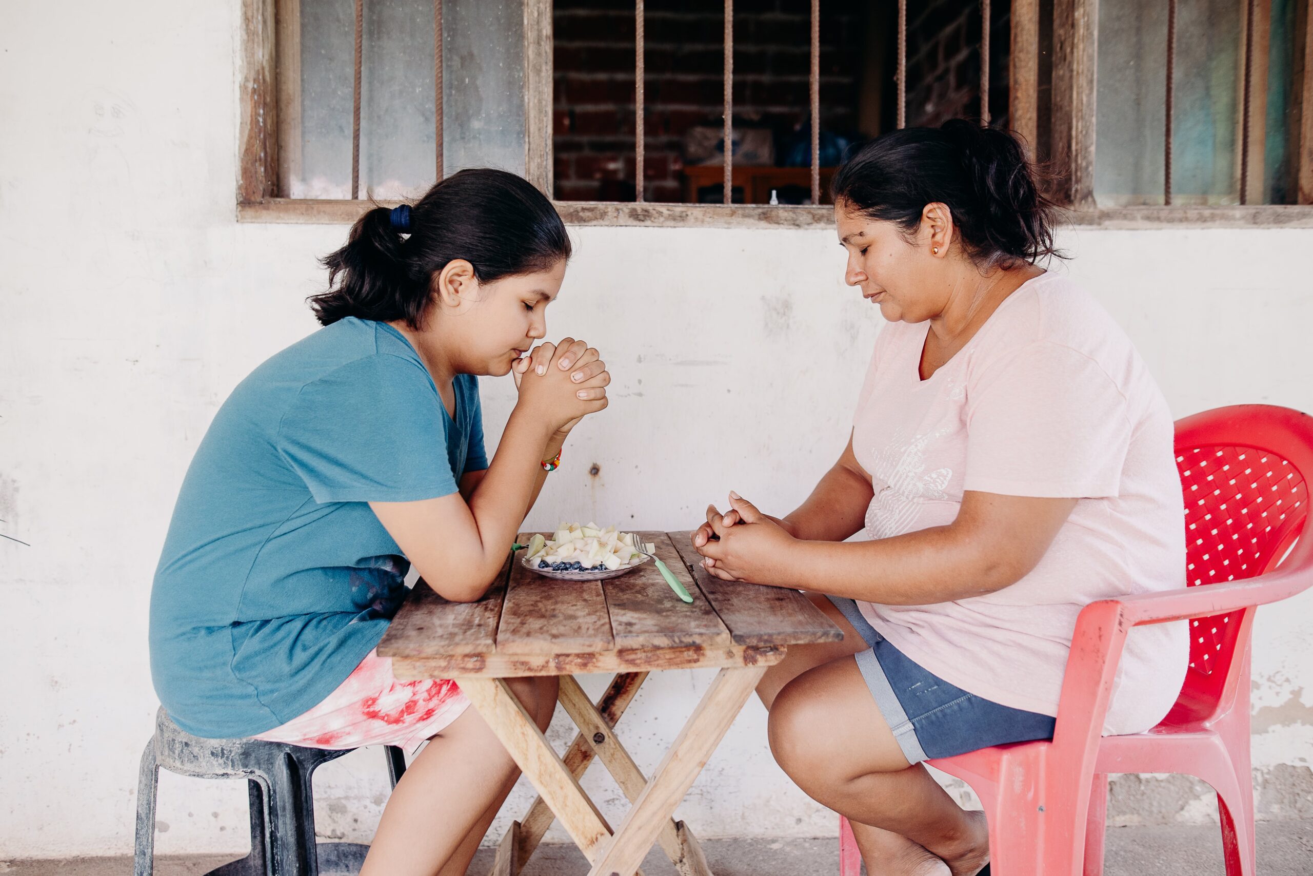 Q2_HB_Feature_Wendy_38-min A young girl and a young woman sit with each other at a wooden table as they clasp their hands and pray.