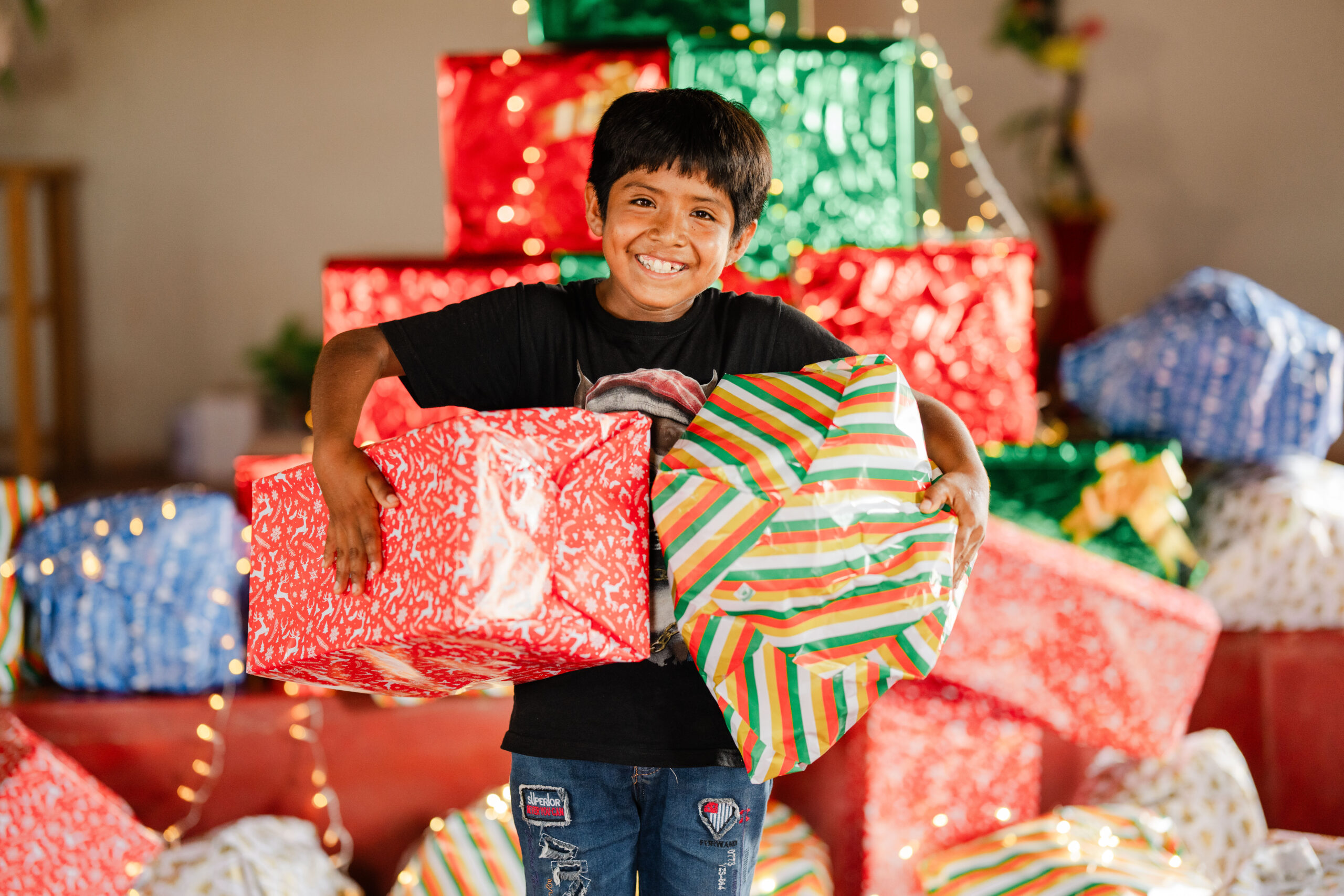 A boy stands in front of a pile of wrapped presents and holds two of them.