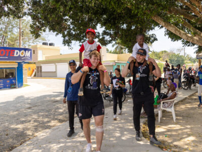 Pro football players carry children on their shoulders down a street in the Dominican Republic. Several children and adults are walking behind them.