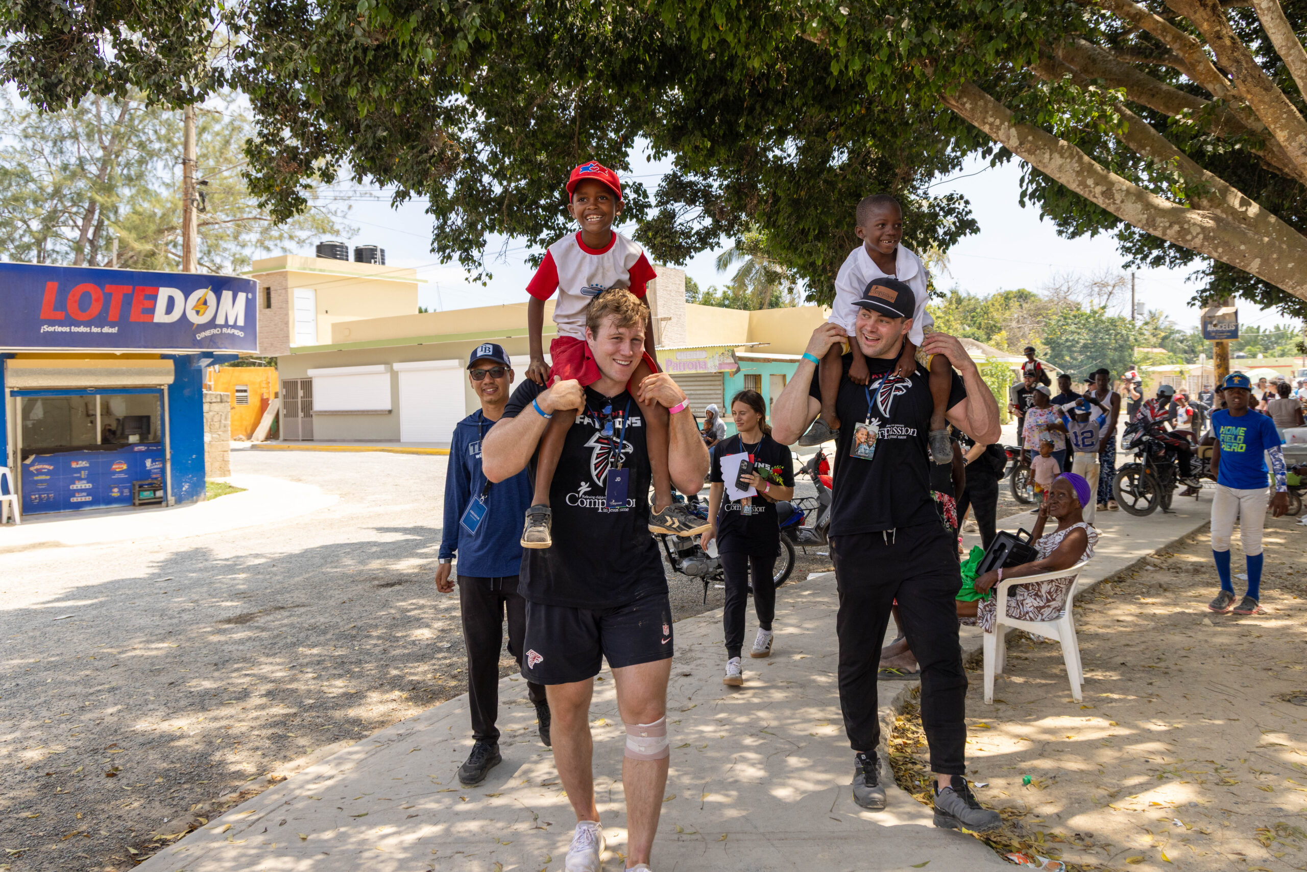 Pro football players carry children on their shoulders down a street in the Dominican Republic. Several children and adults are walking behind them